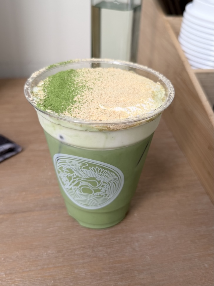An iced green tea matcha latte topped with cream foam, green matcha powder, and kinako roasted soybean powder in a clear cup, matcha einspanner