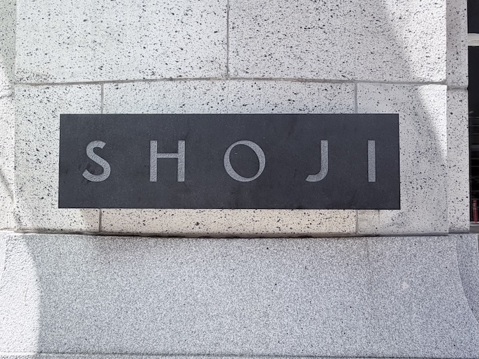 Minimalist black metal sign reading "SHOJI" mounted on a stone wall outside the cafe.