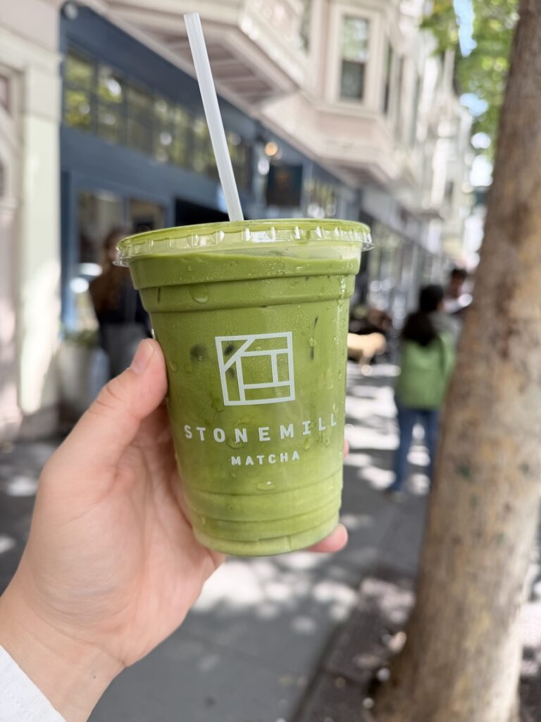 Close-up of a hand holding a cold iced matcha green tea latte in a clear plastic cup with the Stonemill Matcha logo