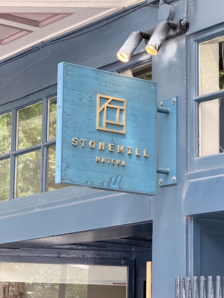 Blue wooden outdoor sign with gold lettering for Stonemill Matcha mounted on a blue cafe facade in San Francisco.