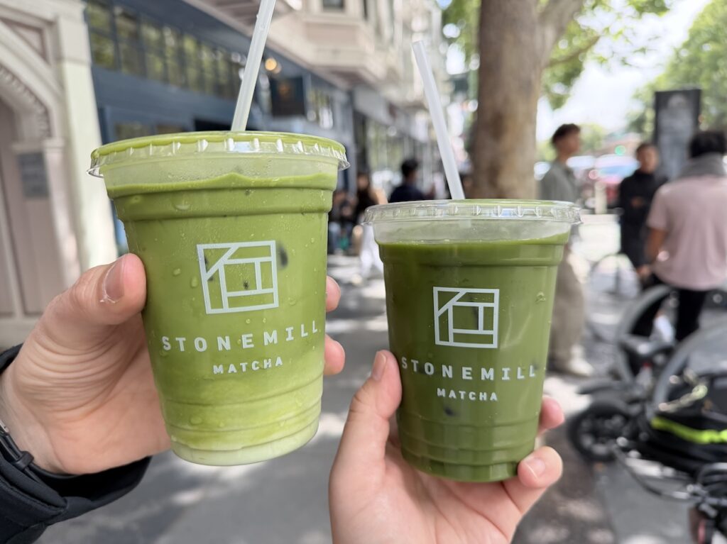 Two people holding cold drinks side-by-side outside Stonemill Matcha, featuring an iced matcha latte and a dark iced green tea.