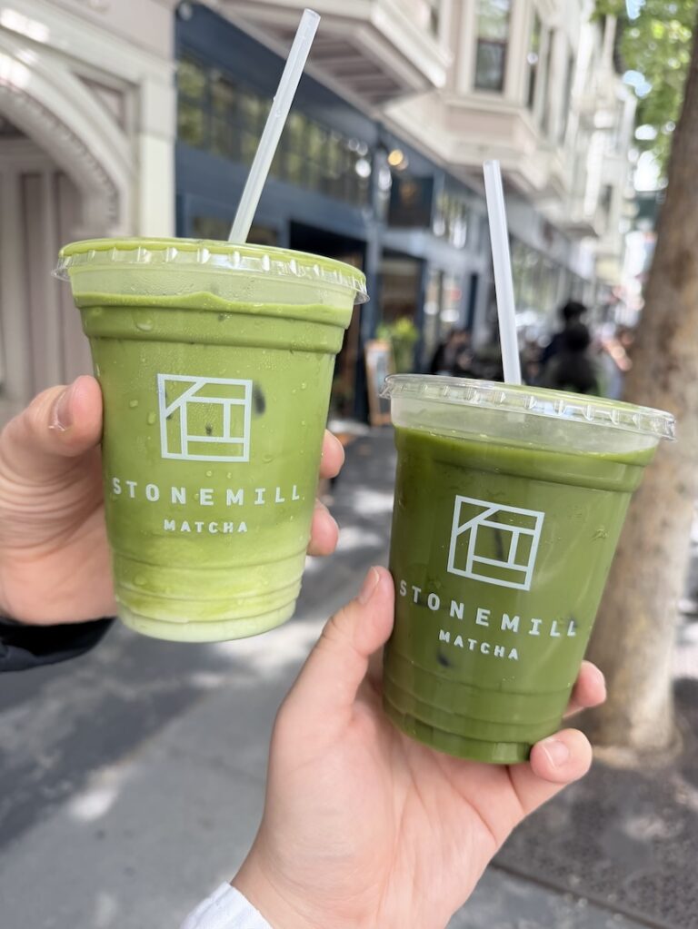 Vertical view of two hands holding iced drinks from Stonemill Matcha cafe in San Francisco, showing a light iced matcha latte and a dark green tea.