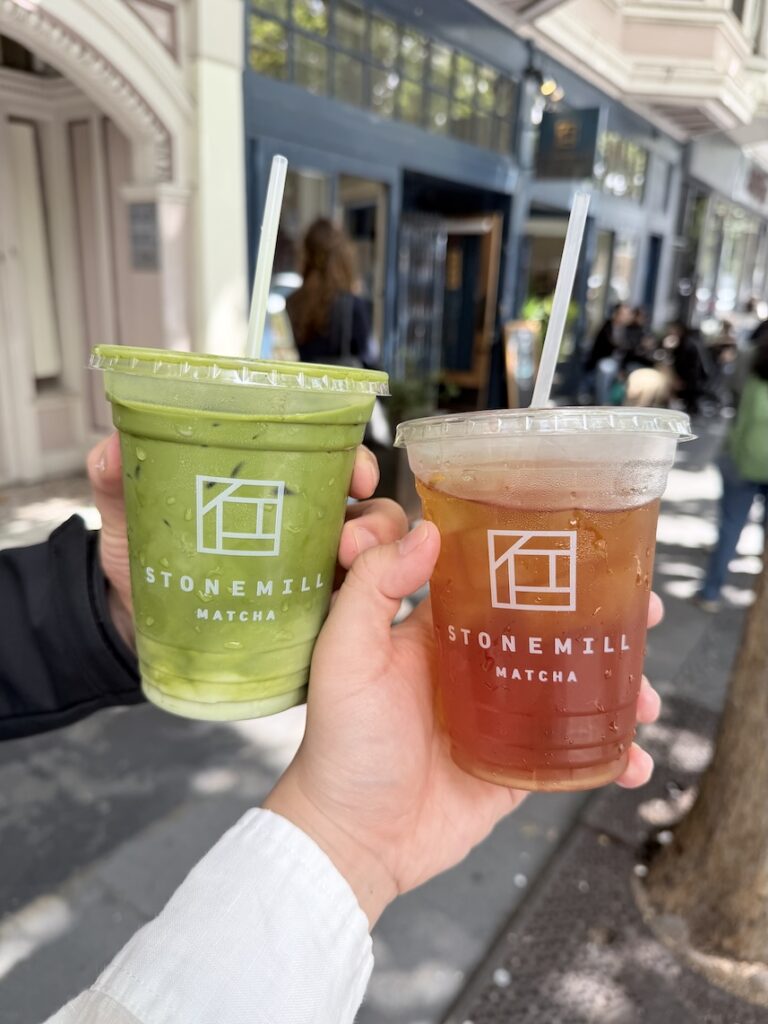 Two hands holding drinks outside Stonemill Matcha, including a creamy iced matcha latte and an iced hojicha roasted green tea.